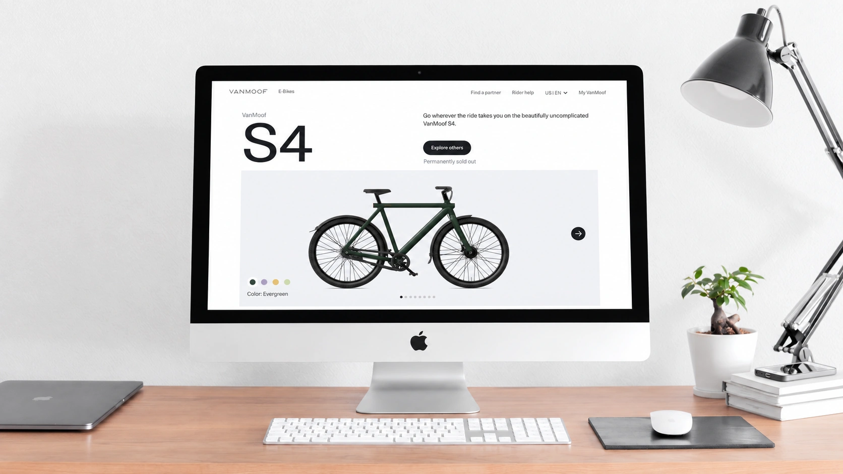VanMoof S4产品页展示在浅木桌面的银色iMac中，旁边是金属台灯与小盆栽